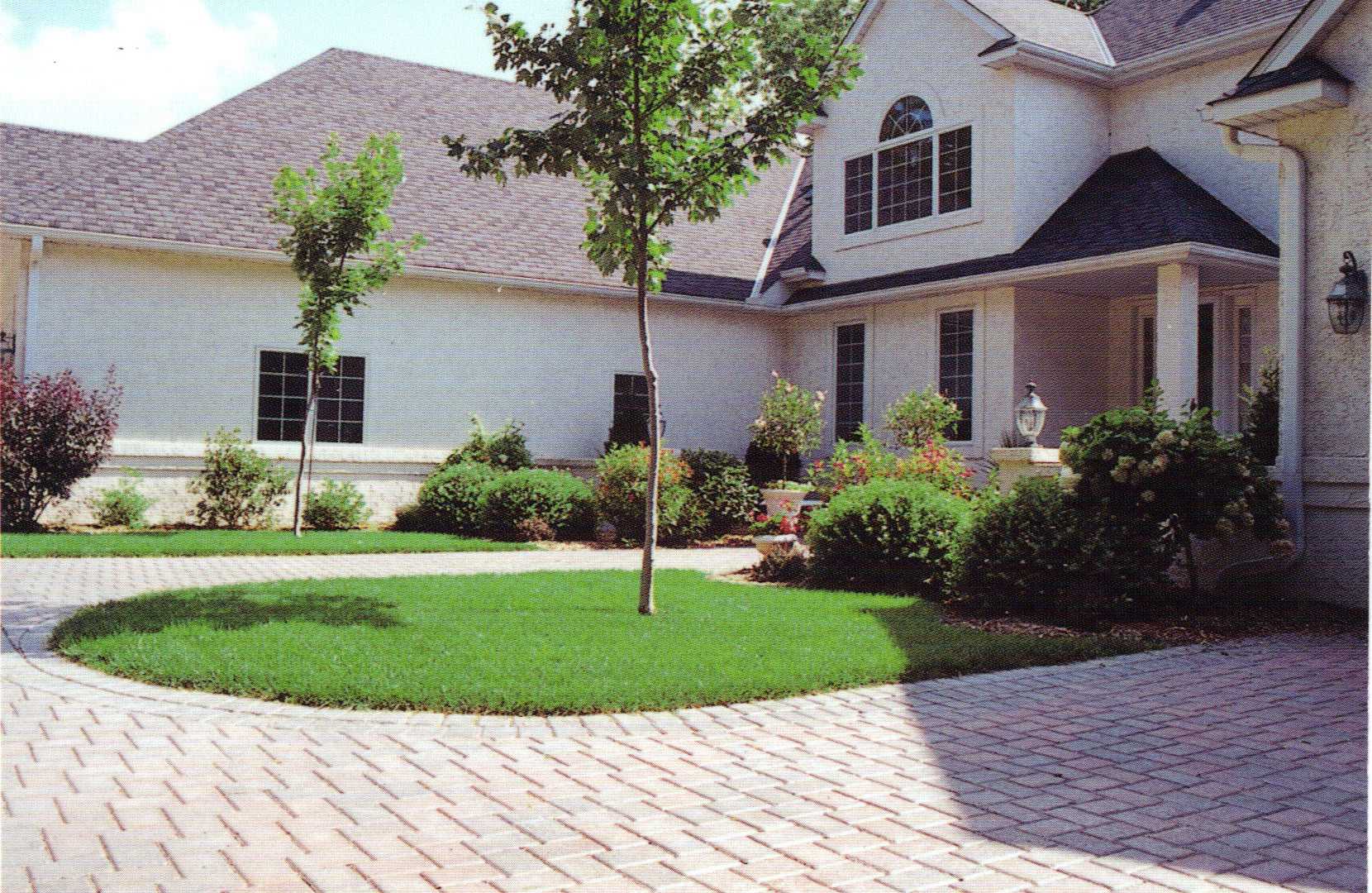 A suburban house with light brick exterior, large windows, and a courtyard featuring a circular lawn, young trees, and well-maintained bushes bordered by a light-colored paver driveway landscaped by Cordes Family Landscaping and Snow LLC.